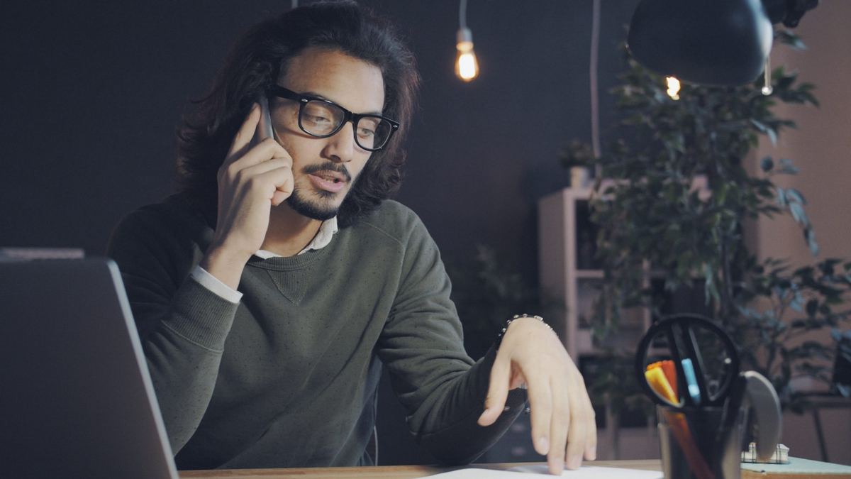 Homem de óculos ao telefone na mesa do escritório, com a mão sobre os papéis ao lado do notebook.